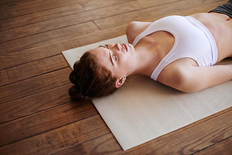 Junge Frau liegt mit geschlossenen Augen und angelegten Armen auf einer Gymnastikmatte.