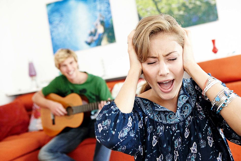 Eine Frau hält sich gestresst die Ohren zu während ein Mann im Hintergrund Gitarre spielt. - Bildnachweis: © wdv / Oana Szekely Eine Frau hält sich gestresst die Ohren zu während ein Mann im Hintergrund Gitarre spielt.