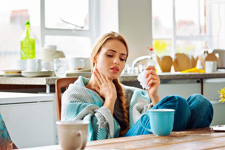 Eine Frau sitzt am Tee trinkend am Küchentisch mit einer Wolldecke um die Schultern und fasst sich an den schmerzenden Hals.