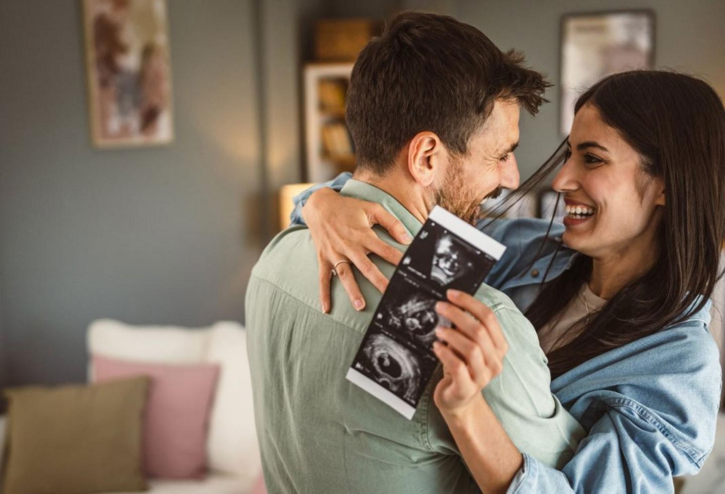 Glückliches Paar umarmt sich, sie hat Ultraschallfots von ihrem Baby in der Hand