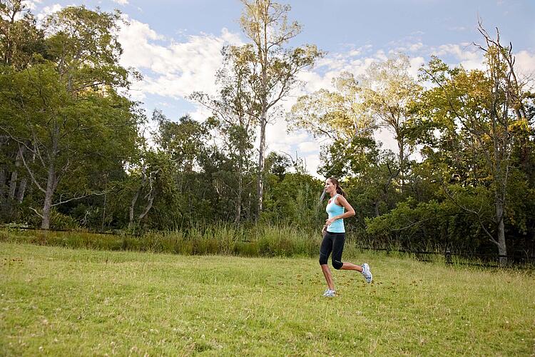 Junge Frau joggt in Sportkleidung über eine Wiese mit Bäumen im Hintergrund.