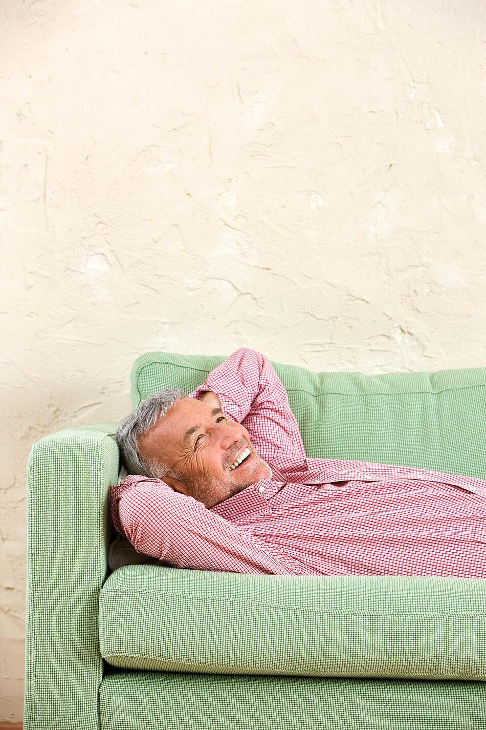 Ein Mann liegt mit hinter dem Kopf verschränkten Armen auf einem grünen Sofa.