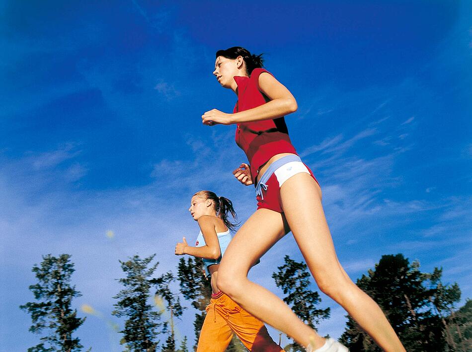 Zwei Frauen in sommerlichem Lauf-Outfit joggen vor blauem Himmel.