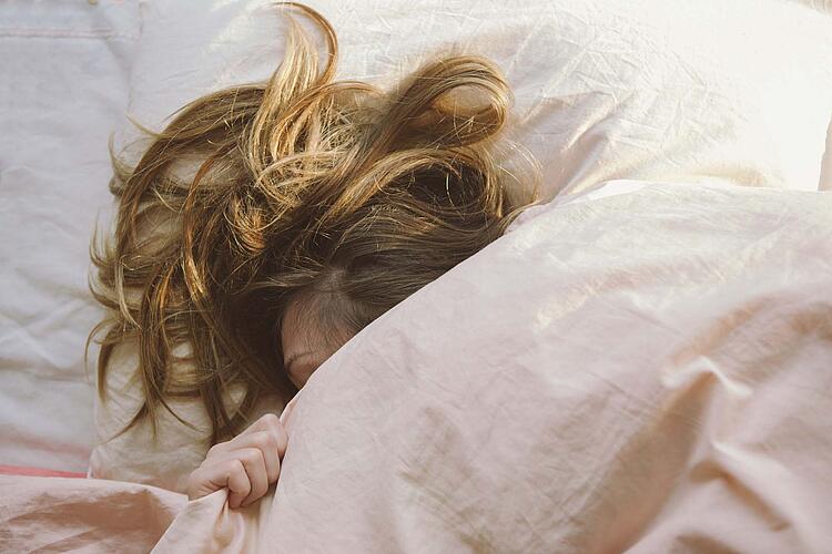 Frau mit langen Haaren liegt im Bett und hat die Decke bis über das Ohr gezogen.