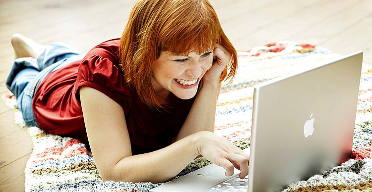 Junge Frau liegt bäuchlings auf dem Boden und tippt lachend auf ihrem Laptop. / Oana Szekely Junge Frau liegt bäuchlings auf dem Boden und tippt lachend auf ihrem Laptop.