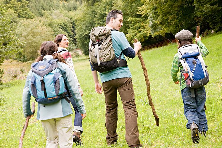 Familie mit Rucksäcken und Wanderstöcken wandert üebr eine Wiese auf einen Wald zu.