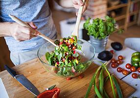 Ein Mann mischt Salat in einer Glasschüssel mit einem Salatbesteck aus Holz.