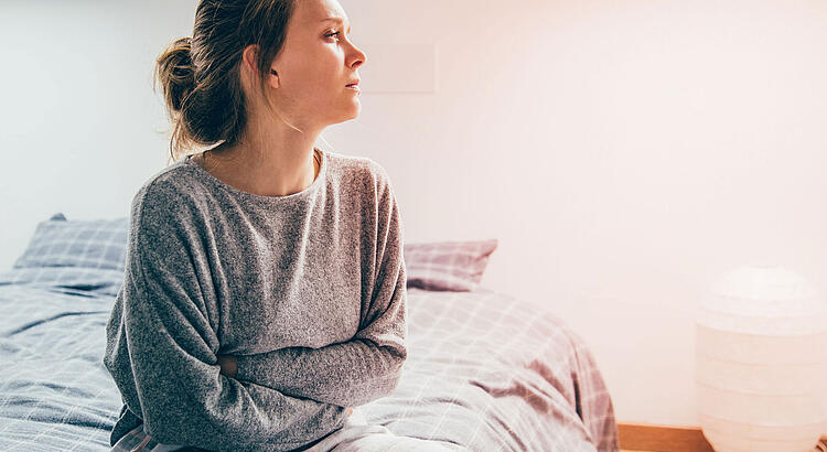 Frau sitzt auf dem Bett und verschränkt die Arme vor dem Bauch.