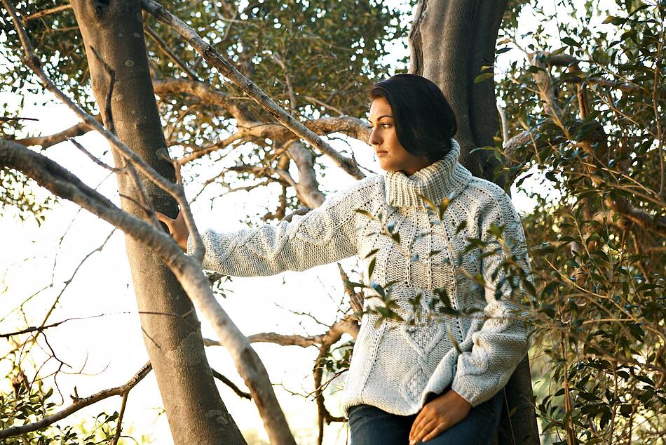 Junge Frau in einem dicken Pullover lehnt an einem Baum.