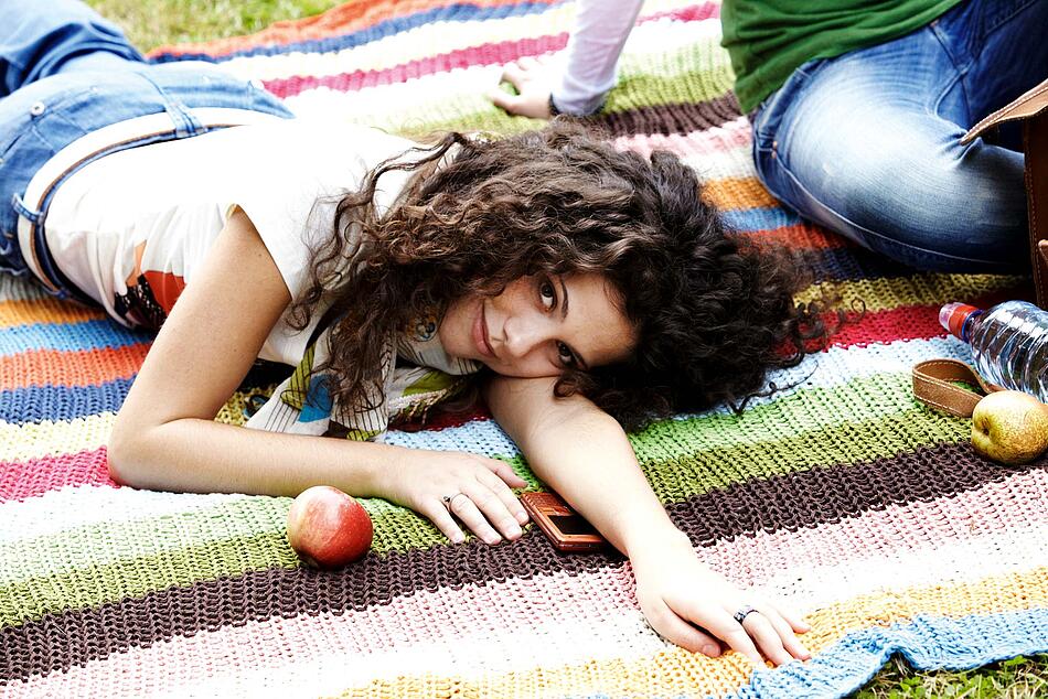 Junge Frau liegt entspannt auf einer Decke, einen Apfel und ein Handy vor sich.