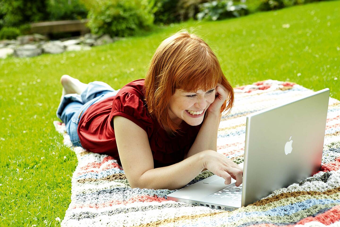 Junge Frau liegt bäuchlings auf einer Wiese und tippt lachend auf ihrem Laptop.
