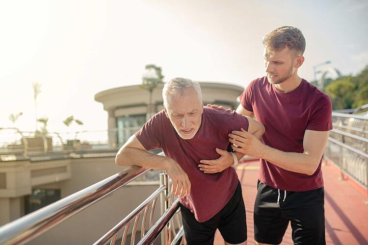 Älterer Mann grefit sich beim Joggen vor Schmerzen ans Herz, junger Mann stützt ihn.