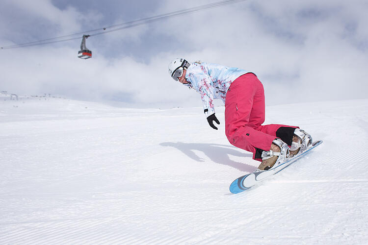 Snowboardfahrer fährt auf einer Skipiste eine Abfahrt hinunter, im Hintergrund ein Sessellift.