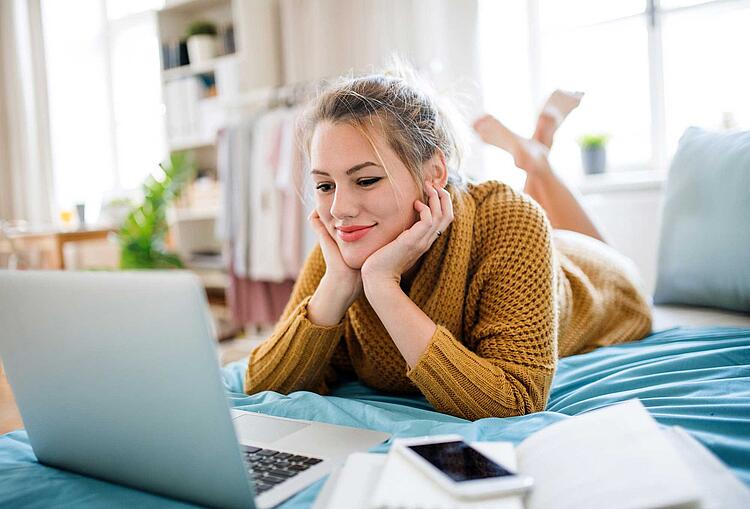 Junge Frau mit Laptop und Smartphone liegt bäuchlings auf dem Bett.