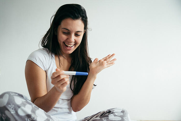 Eine junge Frau schaut lachend auf einen Schwangerschaftstest.