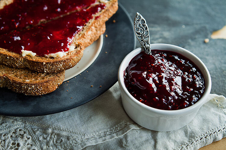 Zwei Scheiben Vollkorntoast mit Himbeer-Marmelade liegen auf einem Teller, daneben ein Schälchen Marmelade mit Silberlöffel.