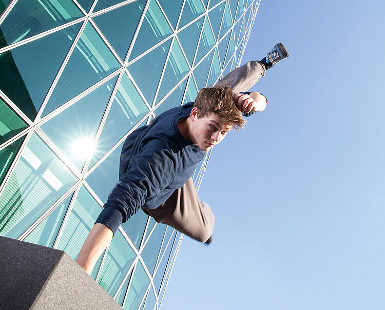 Junger Mann macht Parkour in der Stadt vor einem Gebäude mit Glasfassade.