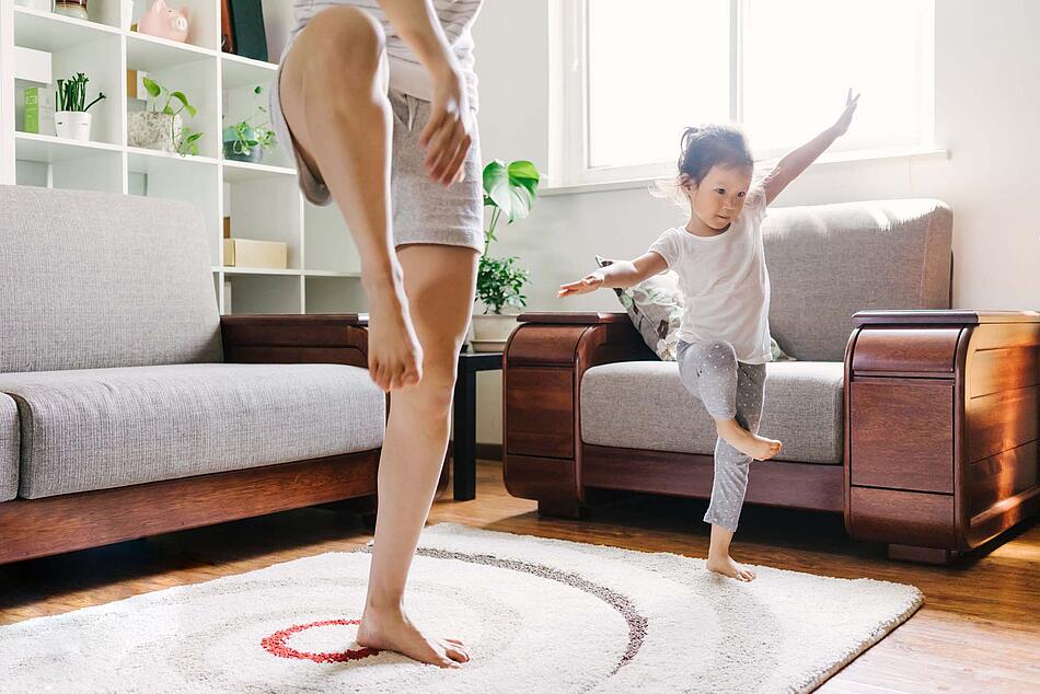 Ein kleines Mädchen macht mit seiner Mutter eine Yogaübung auf dem Wohnzimmerteppich. 