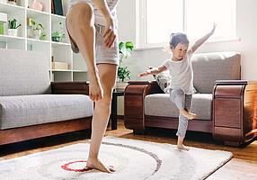 Ein kleines Mädchen macht mit seiner Mutter eine Yogaübung auf dem Wohnzimmerteppich. 