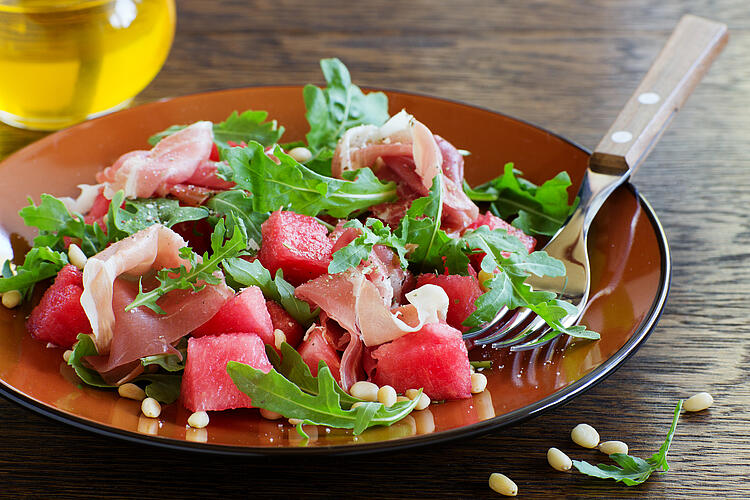 Melonensalat mit Parmaschinken und Rukola auf einem Teller mit Gabel. 