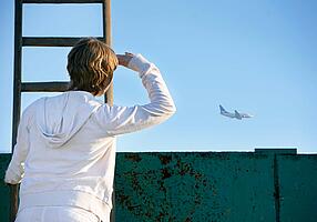 Eine Frau steht auf einer Leiter und beobachtet ein Flugzeug am Horizont.