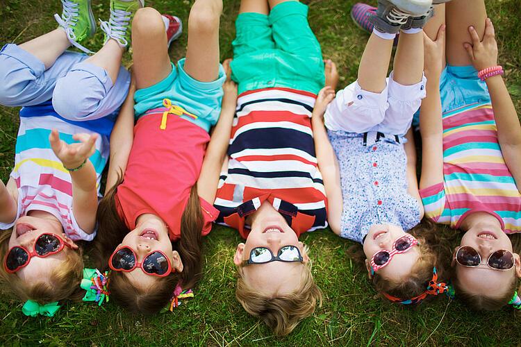 Fünf Kinder liegen auf dem Rücken im Gras tragen Sonnenbrillen und schauen in den Himmel.