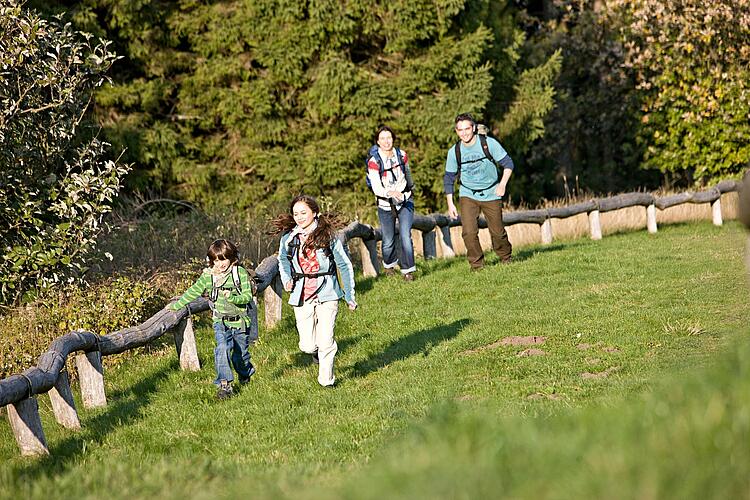 Familie wandert an einem Holzaun entlang über eine Wiese.